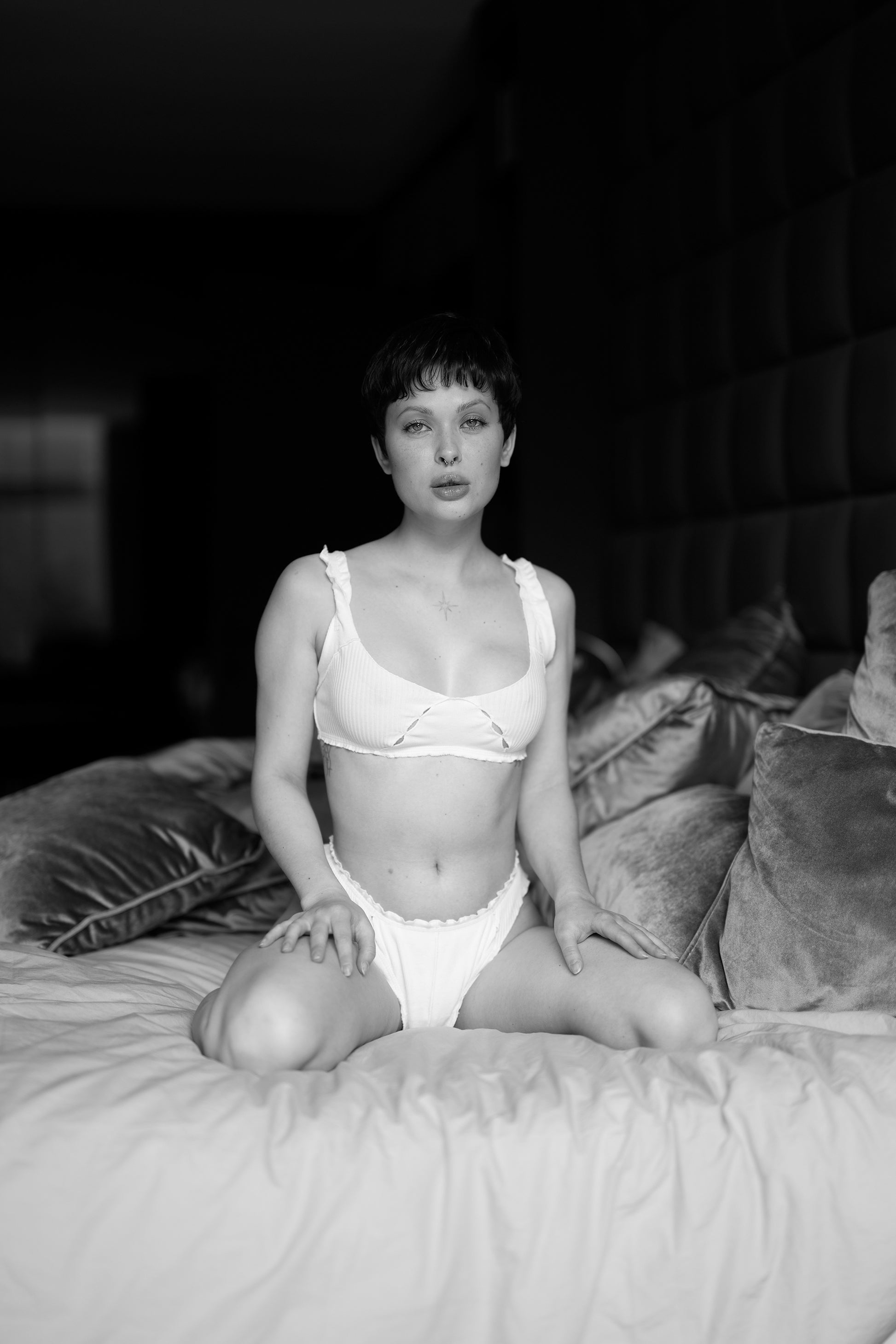 Person wearing a white bra and panties sitting on a bed in a black and white photo.