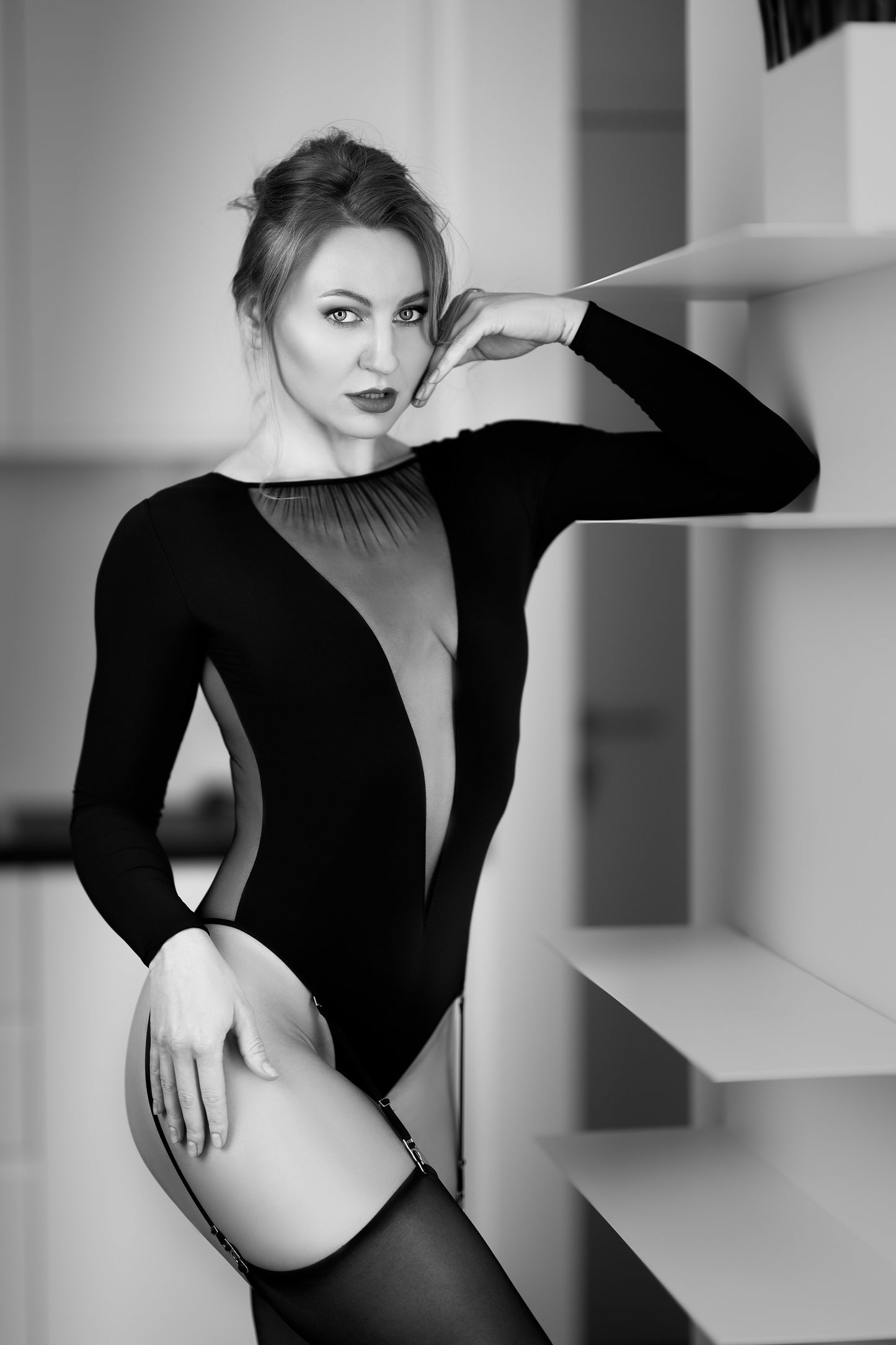 A woman posing in a black long-sleeve bodysuit with a sheer mesh chest panel and thigh-high stockings, set against a modern minimalist gray background with white shelving.